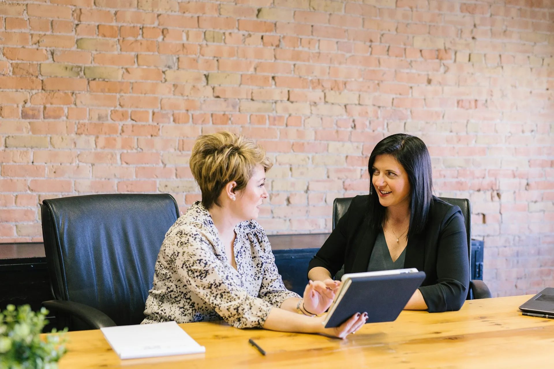 Two professional women discuss work and review information on a tablet during a meeting in a modern office